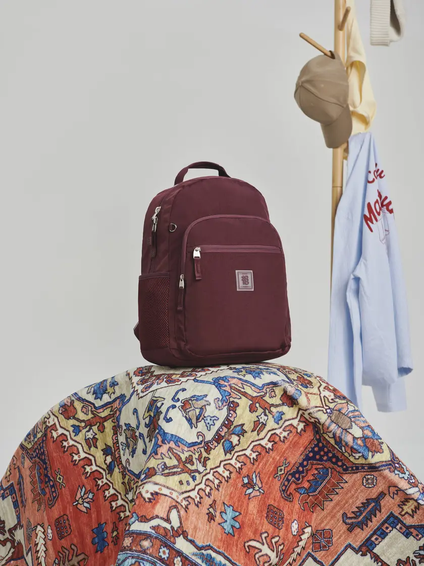 Mochila Chenson color vino apoyada sobre un tapete, junto a un perchero